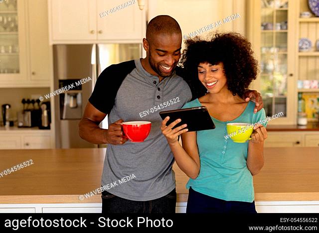 Front view of a mixed race couple standing in the kitchen, holding cups of coffee, embracing, smiling and looking at tablet computer