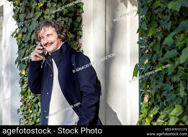 Hombre sonriente hablando por teléfono inteligente fuera de la casa cubierta de hojas de hiedra en un día soleado
