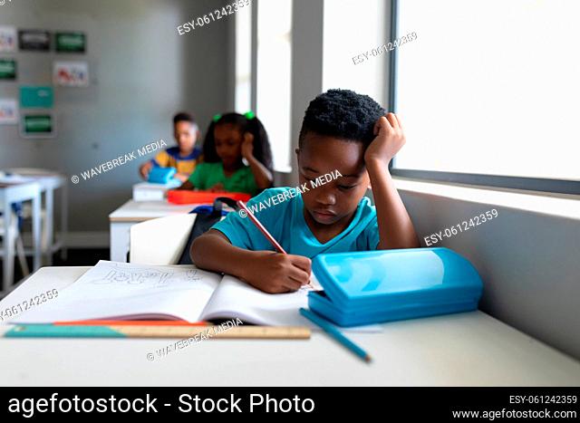 African american elementary schoolboy with head in hand writing on book at desk in classroom