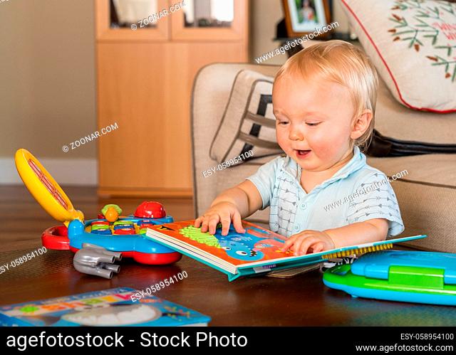 Baby boy sitting on the floor of living room and happily reading a book rather than play with toy computer