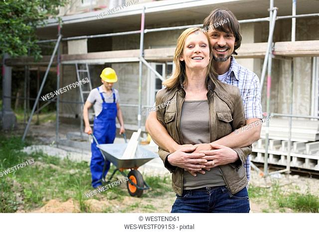 Young couple at site embracing, construction worker in background
