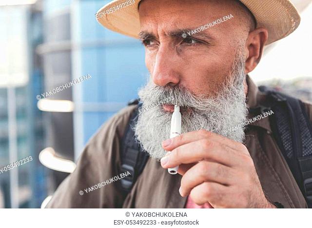 Pensive mature bearded male tourist is smoking electronic cigarette and looking aside with seriousness. He wearing hat. Copy space on left side
