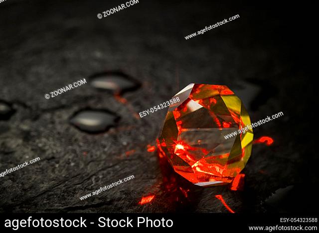 Close-up of a red illuminated crystal with cut and polished facets on a wet slate with individual drops of water. Nahaufnahme eines rot beleuchteten Kristalls...