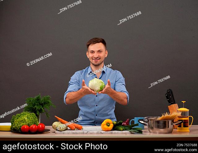 Cute man keeping cabbage in hands. He is going to prepare vegetarian meals in the kitchen. All the necessary products on the counter