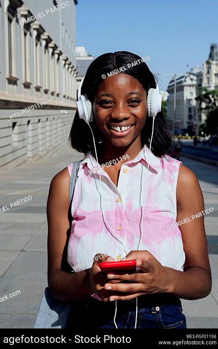 Happy woman with mobile phone listening music through headphones in city on sunny day