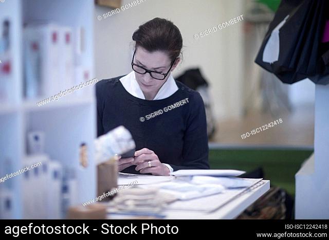 Female tailor wearing glasses, sitting at table, checking her mobile phone