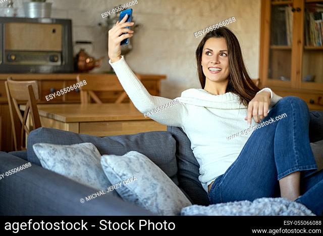 beautiful young smiling woman in white sweater taking selfie on smartphone on gray sofa