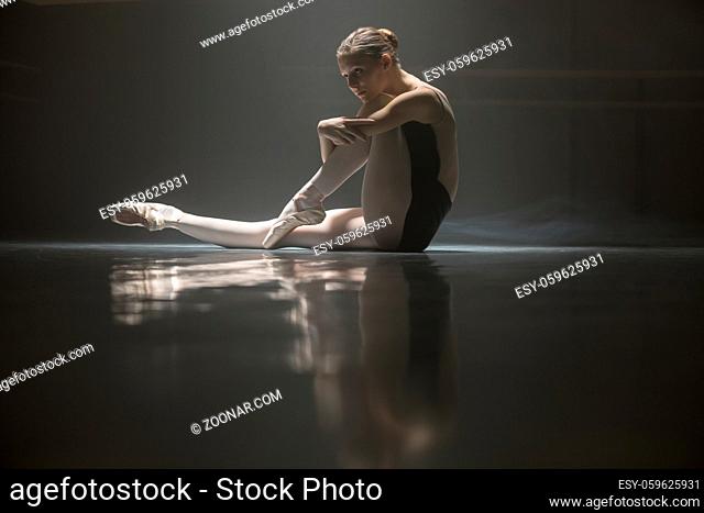 Beautiful ballerina looking in front of her while sitting on the floor in the dance hall. She clasps her hands one leg while the other leg is stretched forward