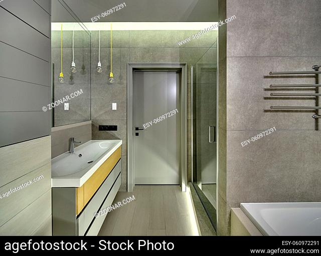 Stylish bathroom in a modern style with tiles on the walls and floor. There is a sink with a faucet, mirror, white bath, glass door, towel holder, entrance door