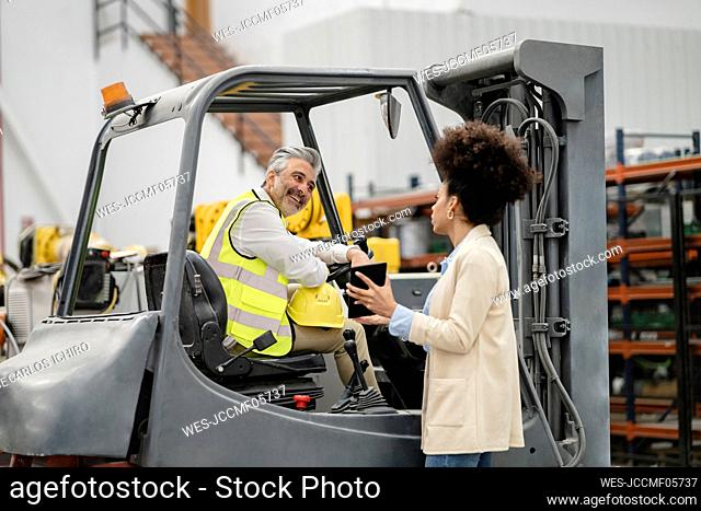 Businesswoman showing tablet PC to engineer sitting in forklift at factory