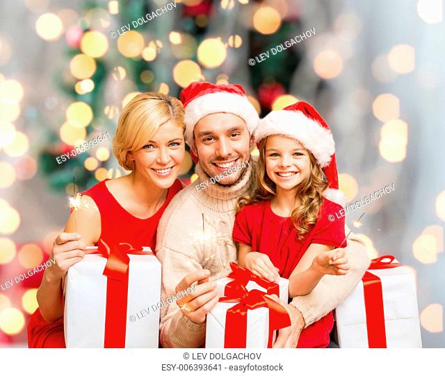 holidays, family and people concept - happy mother, father and little girl in santa helper hats with gift boxes and sparklers over living room and christmas...