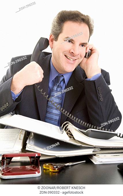 Geschäftsmann am Schreibtisch telefoniert mit seinem Handy und hält mit der anderen Hand den Hörer des Bürotelefons zu