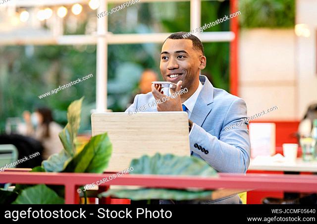 Smiling businessman talking on smart phone through speaker sitting at sidewalk cafe