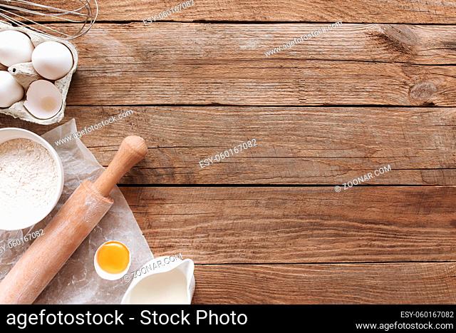 Baking ingredients, kitchen utensils on old wooden background. Cooking dough, preparing egg yolk, flour, rolling pin, milk, parchment paper, whisk, salt, sodium
