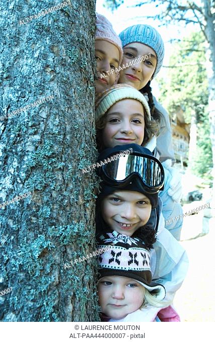 Kids and teens peeking from behind tree in ski clothes, portrait