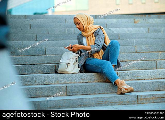 Arab female student sitting on the stairs in downtown. Muslim woman applies makeup at the entrance to the business center. Religion and education