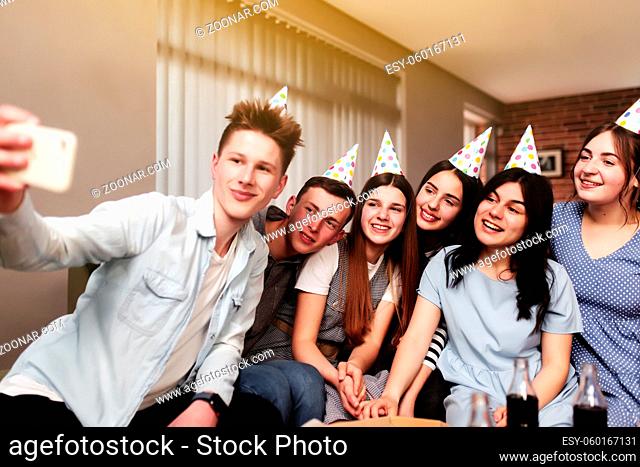 Group of best friends with festive cap taking selfie at birthday party. Girls and guys are posing and smiling at camera of mobile phone