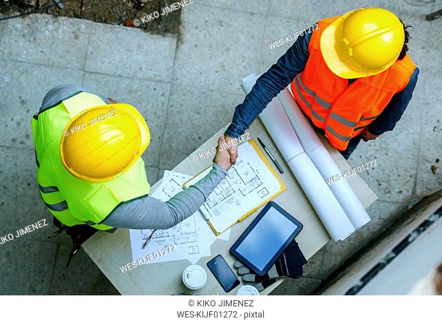 Woman and man in workwear shaking hands above construction plan