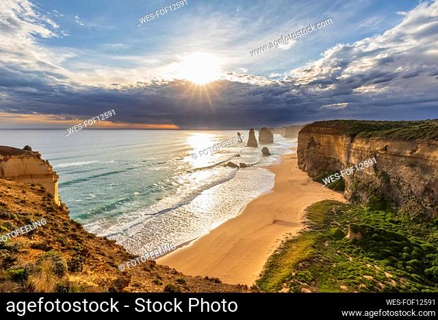 Australia, Victoria, View of sun shining through storm clouds over sandy beach in Port Campbell National Park with Twelve Apostles in background