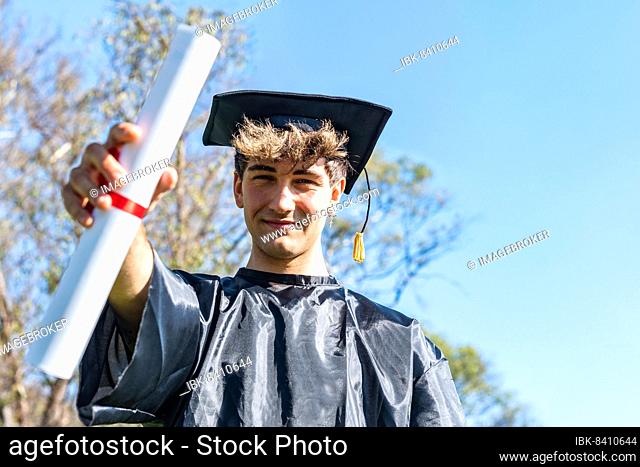 Happy graduated young man wearing a bachelor gown and a black mortarboard and showing his diploma