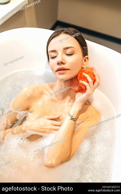 Relaxed lady lying with her eyes shut while washing her body with a soapy nylon loofah