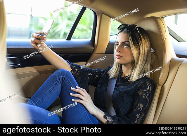 Wealthy and successful young Hispanic woman in back seat of luxury car wearing sunglasses texting on phone