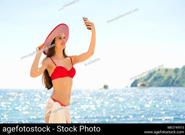 Beautiful young woman wearing a trendy striped hat while posing for a selfie picture on the beach during summer vacation in Flores Island, Indonesia