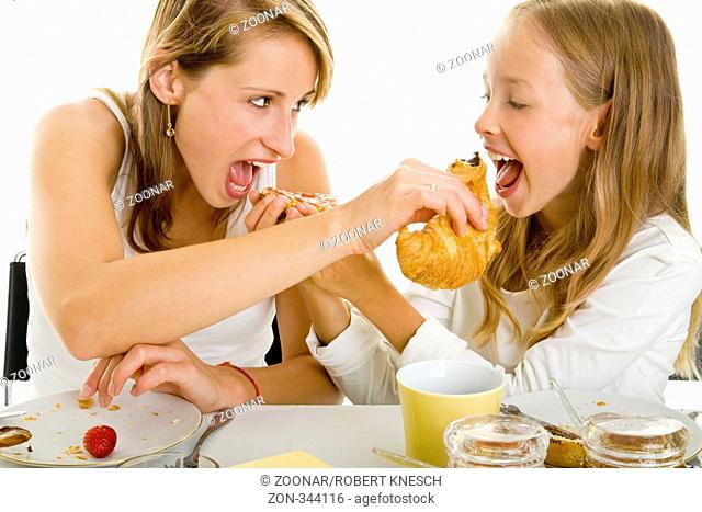 Junge Mutter und ihre Tochter lassen sich am gedeckten Frühstückstisch gegenseitig von ihrem Brötchen und Croissant abbeißen
