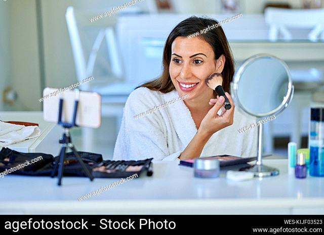 Smiling female influencer vlogging about make-up on smart phone at home
