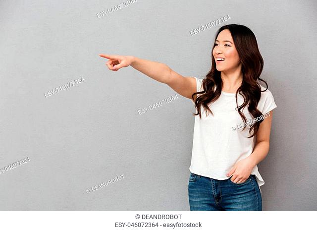 Photo of asian satisfied woman in casual t-shirt and jeans posing on camera and pointing finger aside on copyspace isolated over gray background