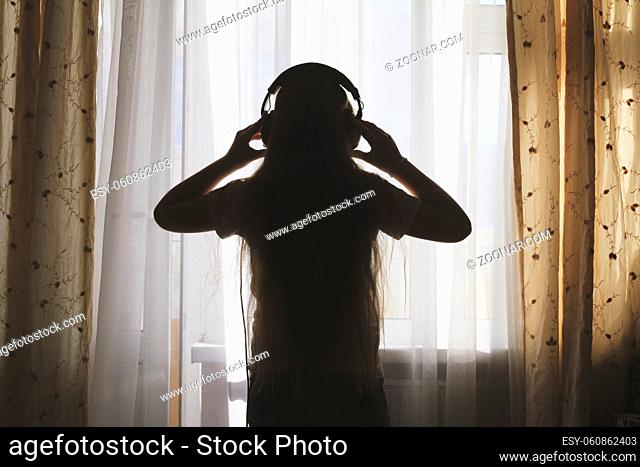 Silhouette of teenager girl in headphones in front of the window, concept of youth leisure