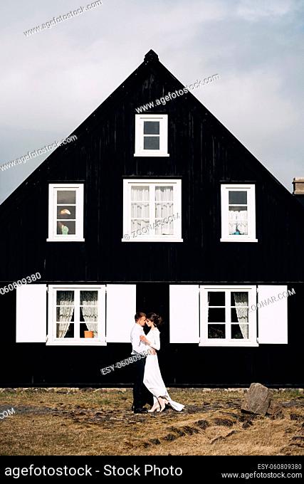 Wedding couple near a black wooden house with white windows and shutters. The groom hugs the bride