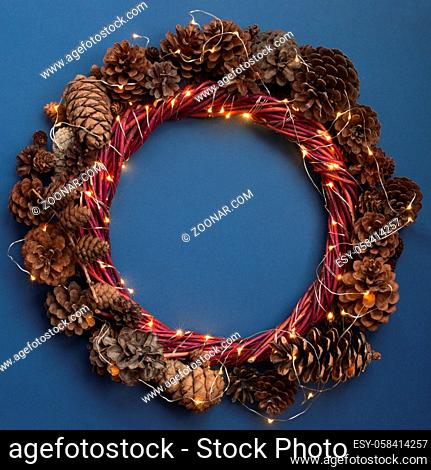 Christmas wreath of pine cones and glowing lights garland on blue background top view flat lay copy space text