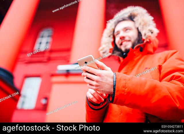 handsome young male student with toothy smile and beard stands on red wall background, facade of educational institution in red winter jacket with hood with fur