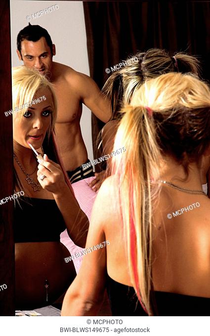 Sexy young blond applying lip gloss in her bedroom with a man standing behind her watching her in the mirror