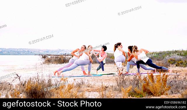 Six attractive women in sportswear doing yoga outdoors, standing on mat in circle on nature performing Side Angle Pose or Parivrtta Parsvakonasana