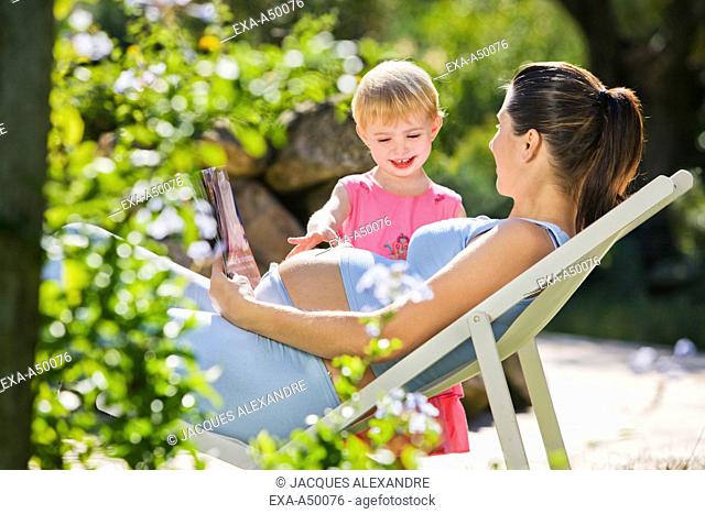 Daughter touching her pregnant mothers belly while she is realxing in the garden on a lounge chair
