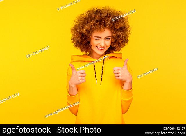 Good job. Portrait of optimistic curly-haired young woman in urban style hoodie winking and showing thumbs up, approval gesture