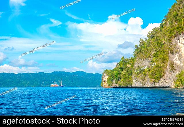 The rocky coast of the tropical island of Indonesia in sunny weather. Rainforest on the slope. Sailing yacht in the distance