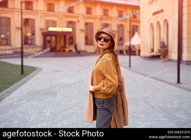 Turned from behind fashion model young woman in sunglasses and hanging on a shoulder autumn beige coat walking on the street happily posing for the camera