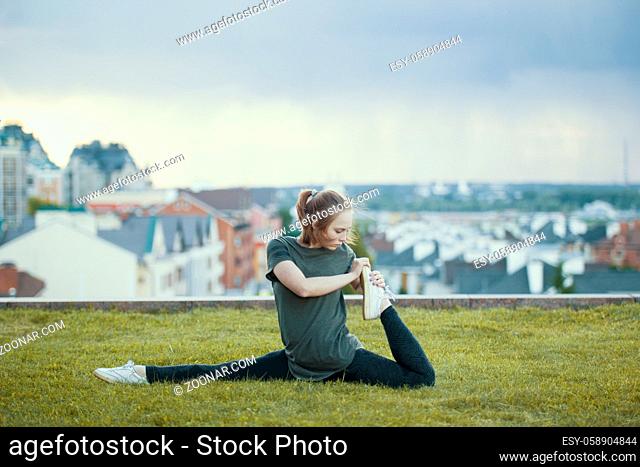 Young female acrobat on the grass performs twine, cityscape on background, horizontal
