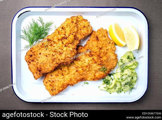 Traditional deep fried Wiener schnitzel from veal topside with cucumber salad and lemon slices offered as top view on a white tray