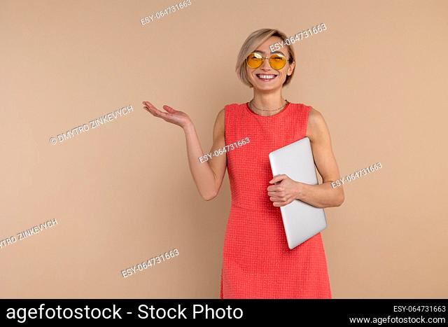 Smiling beautiful woman in dress and sunglasses holding laptop copy space isolated over beige background