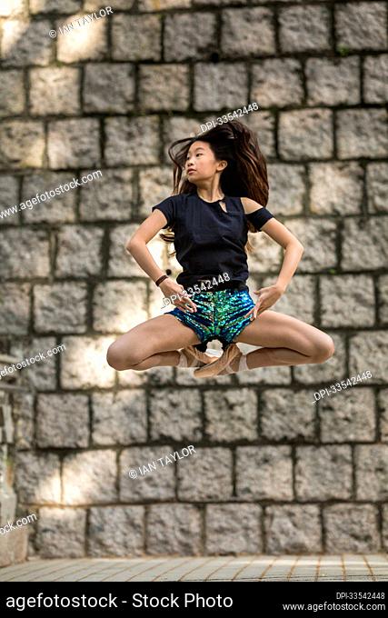 Girl dancing with ballet shoes and in a pose mid-air against a stone wall at a public park; Hong Kong, China