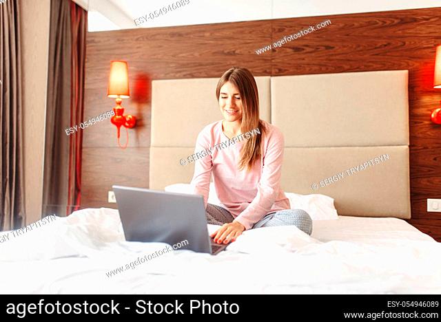Smiling woman sitting in bed and uses laptop, good morning, bedroom interoir on background