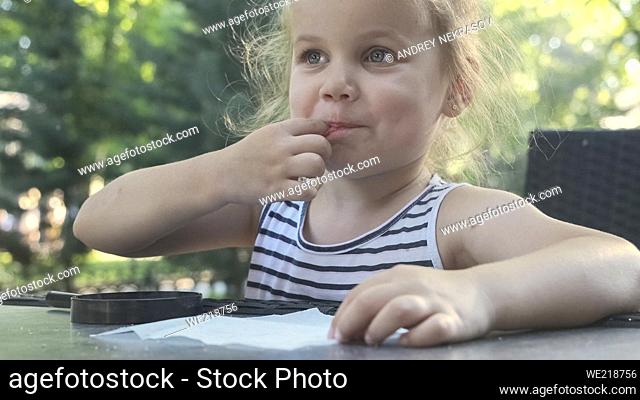 Little girl tastes salt. Close-up portrait of blonde girl takes salt from napkin with her finger and tastes it while sitting in street cafe on the park
