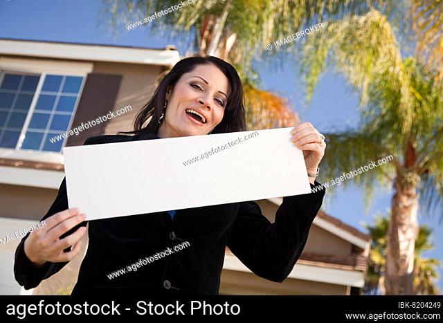 Happy attractive hispanic woman holding blank sign in front of house