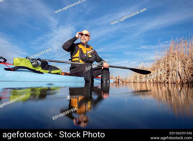 senior male paddler is relaxing in the afternoon sun after workout on a stand up paddleboard, lake in Colorado, winter or early spring scenery, recreation