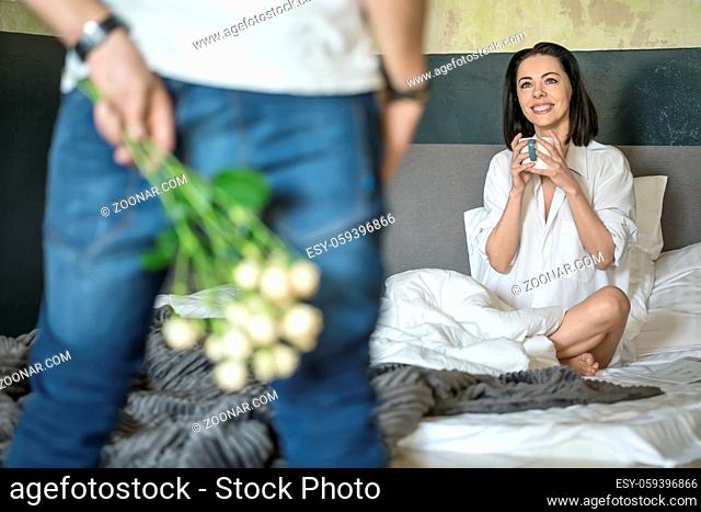 Man in blue jeans and white T-shirt holds a bouquet of roses behind his back. He stands opposite his smiling girlfriend in a white shirt who sits on the bed...