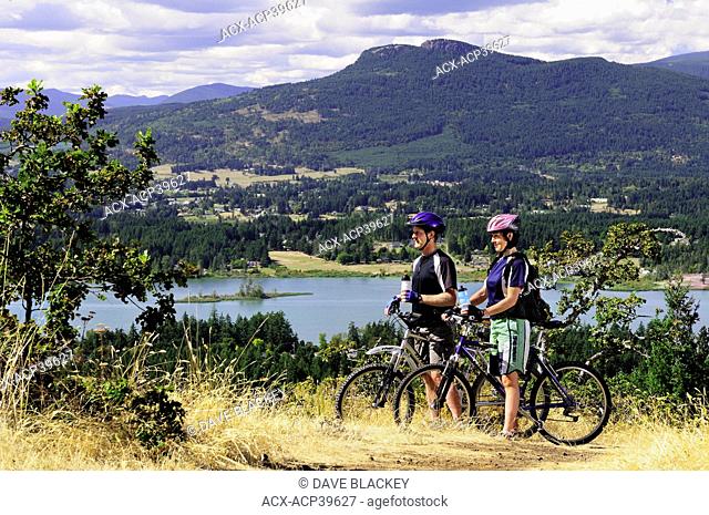 Couple taking a break and enjoying the views while mountain biking on Mt. Tzouhalem with Mt. Prevost in the background in Duncan, British Columbia, Canada